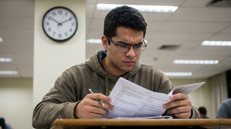 Um jovem de óculos e moletom marrom está sentado em uma sala de aula, segurando e examinando atentamente uma folha de prova. Há um relógio de parede redondo fixado na coluna ao fundo