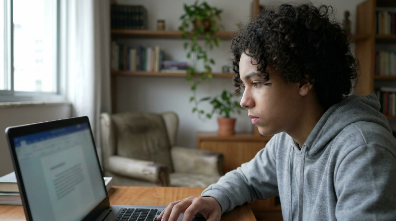 Um jovem com cabelo encaracolado e camisola com capuz cinzenta está sentado a olhar atentamente para o ecrã de um computador portátil numa sala bem iluminada com estantes de livros ao fundo