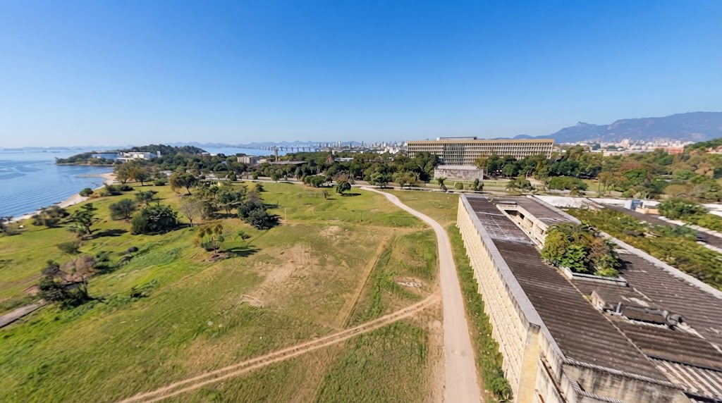 Vista aérea panorâmica de uma zona costeira com extensas áreas verdes, caminhos de terra batida, edifícios de arquitetura em betão e o mar ao fundo sob um céu limpo
