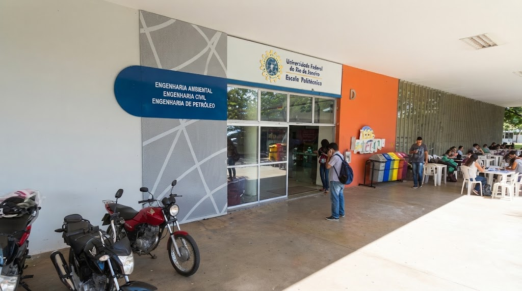 Entrada exterior de um edifício universitário com letreiros azuis indicando cursos de engenharia, com duas motas estacionadas no exterior e estudantes reunidos em mesas ao ar livre
