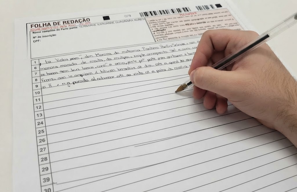 Um plano de pormenor focado numa mão a escrever com uma esferográfica preta sobre uma folha de redação pautada e numerada, com linhas de texto manuscrito já preenchidas nas primeiras linhas