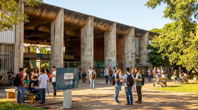 Um grupo de jovens estudantes universitários em pé, conversando e interagindo em uma praça do campus da Universidade de Brasília (UnB). Ao fundo, imponentes edifícios de concreto e árvores sob céu azul. O letreiro