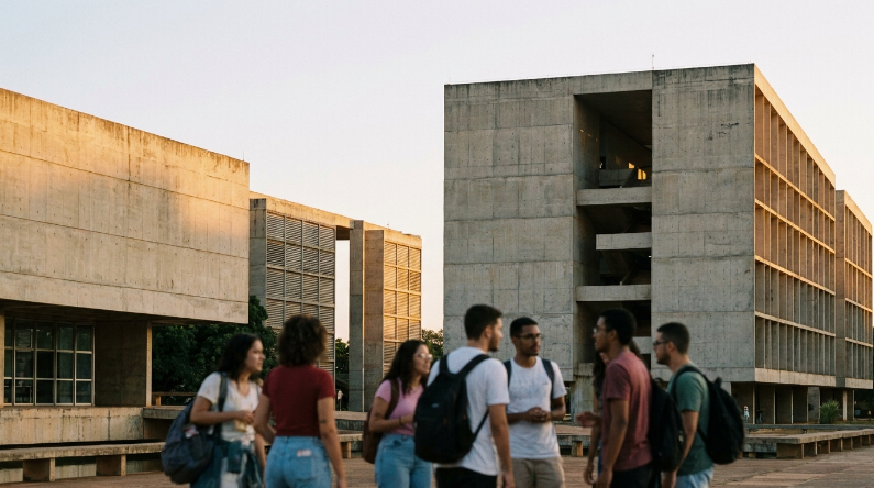Grupo de estudantes universitários em pé, conversando em uma praça ao ar livre em frente a imponentes edifícios de concreto com design arquitetônico marcante
